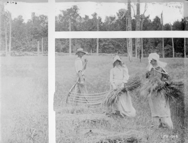 One man and two women are posing standing while working in a field. The women are wearing bonnets and are holding bundles of grain in their arms, and the man is holding a cradle scythe.