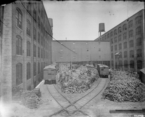 Elevated view of a factory yard. There are brick factory buildings on the left and right, connected by an elevated walkway. There are water towers on the roofs of the factory buildings. In the yard are two sets of railroad tracks, which cross in the foreground. There are railroad cars on the tracks on the left and right, and piles of wood are stacked in between.