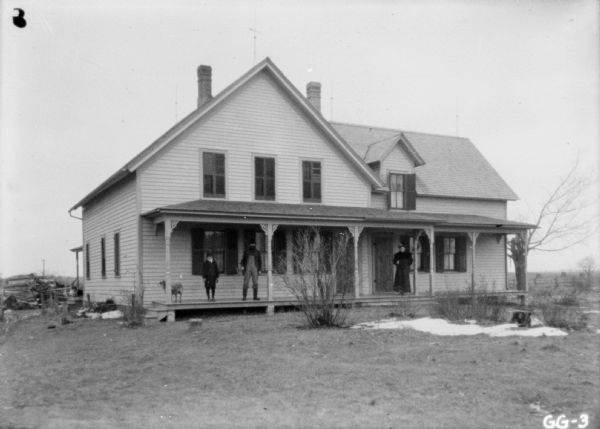 View across lawn towards a family of a man, woman and child, including a dog, posing on the porch of a large house.