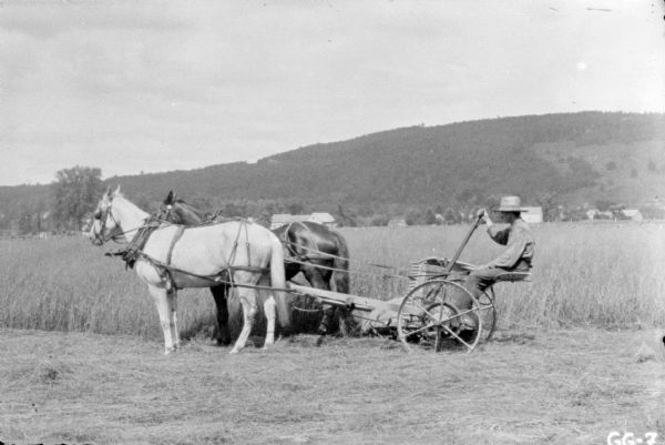 Left side profile view of a man sitting on a horse-drawn mower. Buildings are in the background in the distance at the edge of the field, and a tree-lined hill is in the far background.