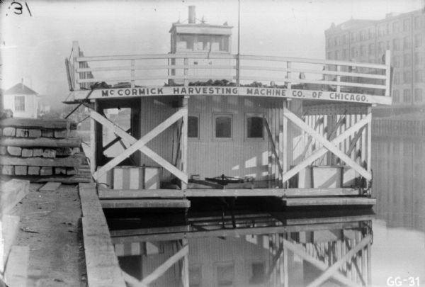 View along dock towards a boat with two decks (float?) anchored in harbor at McCormick Works. Painted on the back of the boat is a sign that reads: "McCormick Harvesting Machine Co. of Chicago."