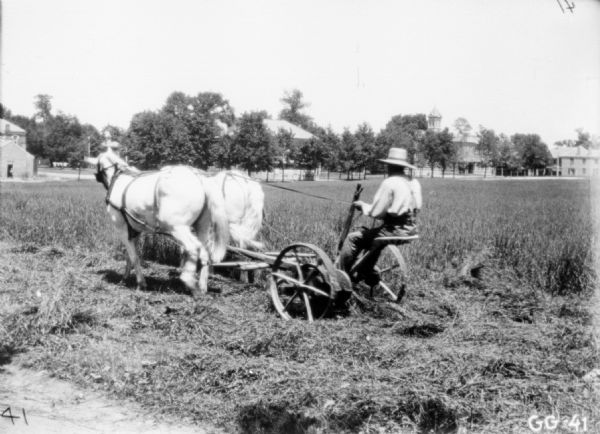 Three-quarter view from left rear of a man using a horse-drawn mower in a field. Buildings are in the background.