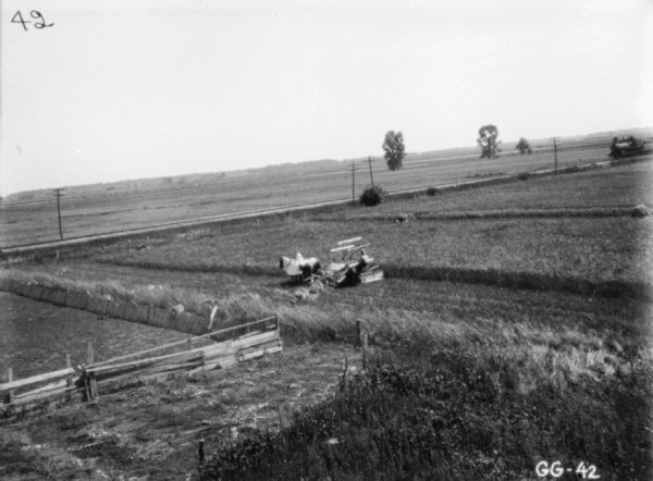 Elevated view of a man using a horse-drawn binder in a field. The horses are wearing blankets. Railroad tracks are running along the field, and a locomotive is on the right coming down the tracks.