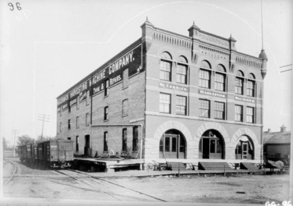 View across road and railroad tracks towards a brick dealership building. Railroad cars are parked along the loading dock. Along the top of the building is a painted sign that reads: "McCormick Harvesting Machine Company."