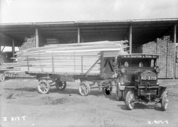 Truck with trailer loaded with lumber. More lumber is stacked under a roofed storage area in the background. Sign on front and side of truck reads: "W.R. Barton & Son, LTD, Timber Merchants."