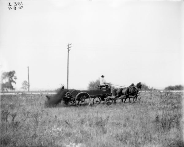 View across field towards a man using a horse-drawn manure spreader. There is a road running along in the background with power line poles.