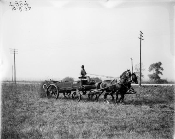 View across field towards a man using a horse-drawn manure spreader. There is a road running along in the background with power line poles.