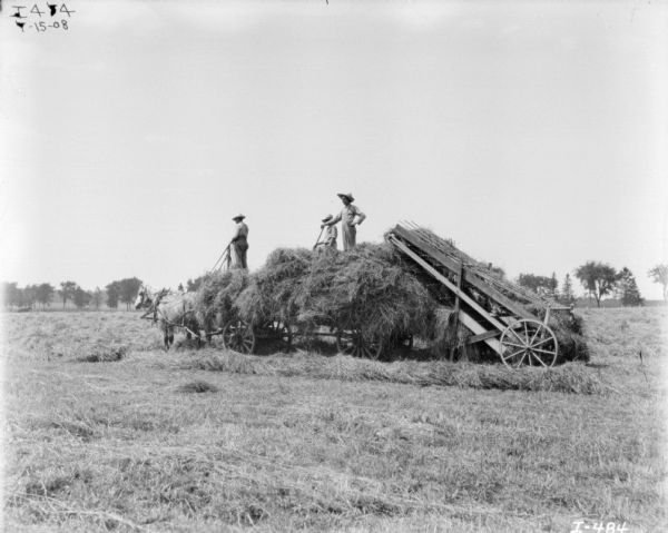 View across field towards men standing on a horse-drawn wagon piled with hay. A hay loader is on the back of the wagon.