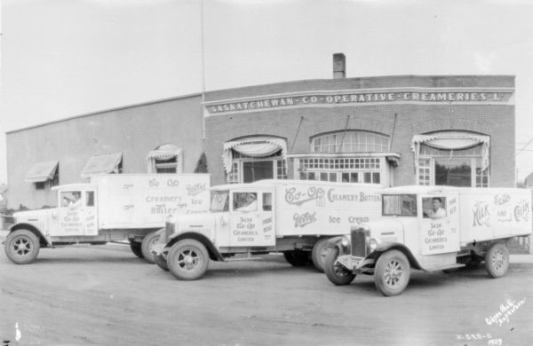 Three men are sitting in delivery trucks in front of a building. The building has a sign that reads: "Saskatchewan-Co-Operative Creameries Ltd."