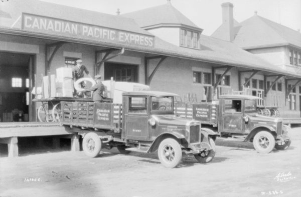 One man is standing on a cart on the loading dock handing another man packages to load into a truck backed up to the loading dock. Another truck is parked on the right. Both trucks have a sign on the side of the bed that reads: "Canadian Pacific Express."