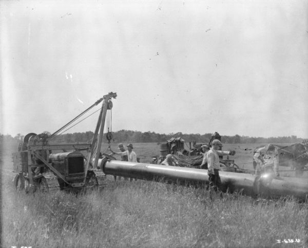 Men laying pipe in a field. An industrial tractor on the left has a pulley to lower the pipe. Behind the men is a horse-drawn wagon and another piece of machinery. One of the men appears to be cutting the pipe with a torch.