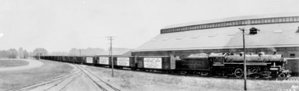 Panoramic view across railroad tracks towards a train with signs on the cars that read: "Train Load of International Trucks for Dealers Philadelphia Territory." Large buildings are behind the train.