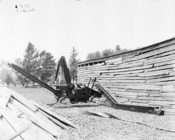 Separator, unfolded, set up outdoors near a farm building. A belt is stretched out on the ground near the separator, perhaps for driving the machinery.
