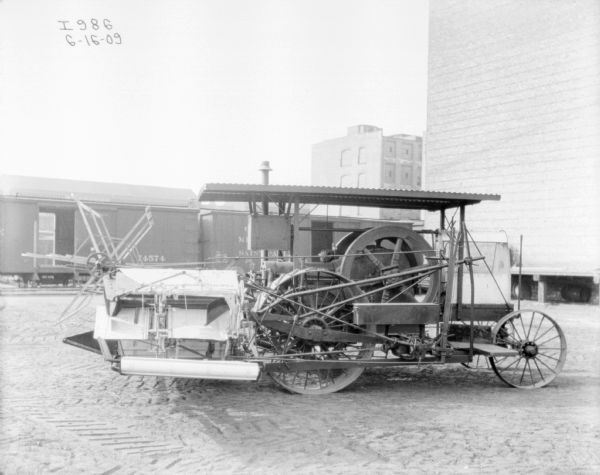 Tractor drawn binder parked outdoors. Railroad cars are in the background, and large buildings are on the right.