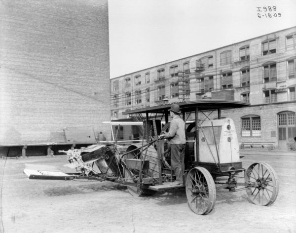 Man standing on tractor drawn binder in a factory yard. In the background are buildings.