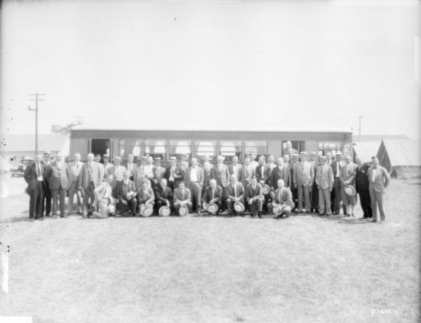 Large group of men wearing suits posing in front of a railroad car outdoors. There are tents in the background on the right.