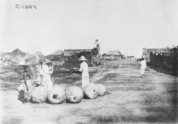 Men are standing in a road with large spools of hemp being wound for transport. Buildings are in the background.