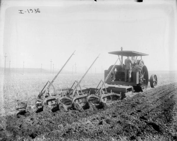 Three-quarter view from right rear of a man using a tractor to plow a field. A line of power poles are in the background on the left.