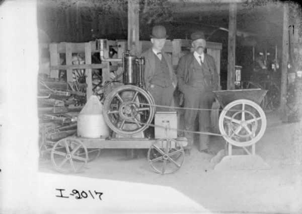 Two men are posing inside a shed beside engine-powered feed mill.