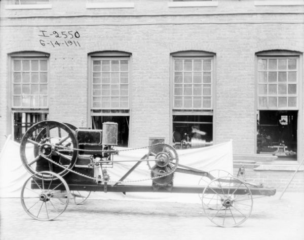 Side view of prototype parked in front of a brick factory buildings. A sheet is hung below large open windows in the background. Through the center window a man can be seen working with a piece of machinery.