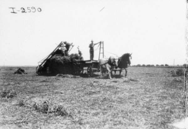 View across field towards a man standing on a horse-drawn wagon, and two men standing on a haystack. They are working with a hay loader.