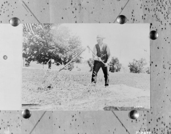Man using a scythe in a field. In the background is a tree, and in the upper left corner is an American Flag.