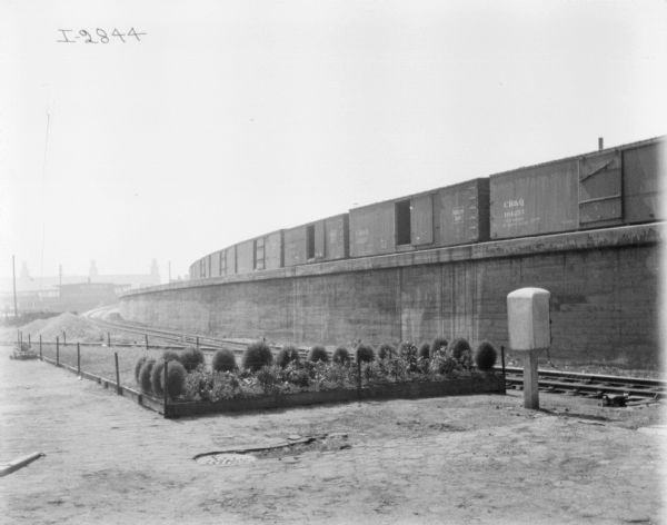 Fenced gardens near railroad tracks. Railroad cars are on another elevated set of tracks. Buildings are in the far background.