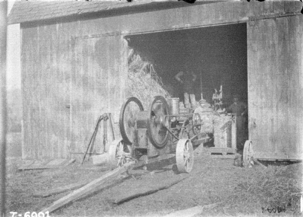 Powered hay press set up in an open barn door. Three men are standing inside near the machinery.
