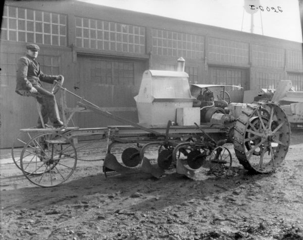 Man sitting on an experimental machine with plows. Factory buildings are in the background.