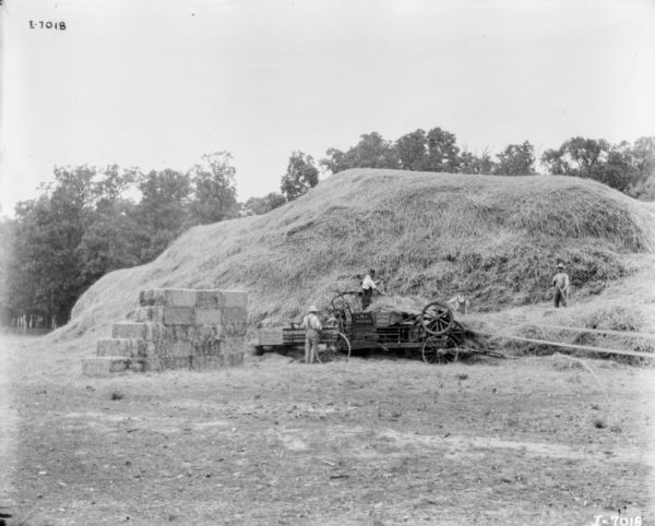 Men and a young boy standing on and near a powered hay press set up in a field. A large haystack is behind them, and hay bales are stacked on the left.