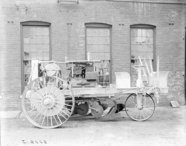 Right side view of tractor parked in front of brick factory building in plant yard. Plow is attached underneath.