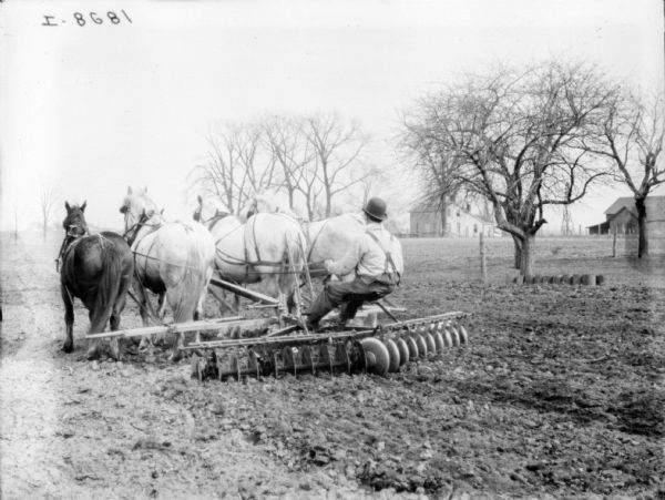 Three-quarter view from left rear of a man using a horse-drawn disk harrow in a field. Farm buildings are in the background on the right.