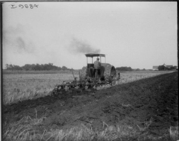Three-quarter view from left rear of a man using a tractor to plow a field. Farm buildings are in the background on the right.