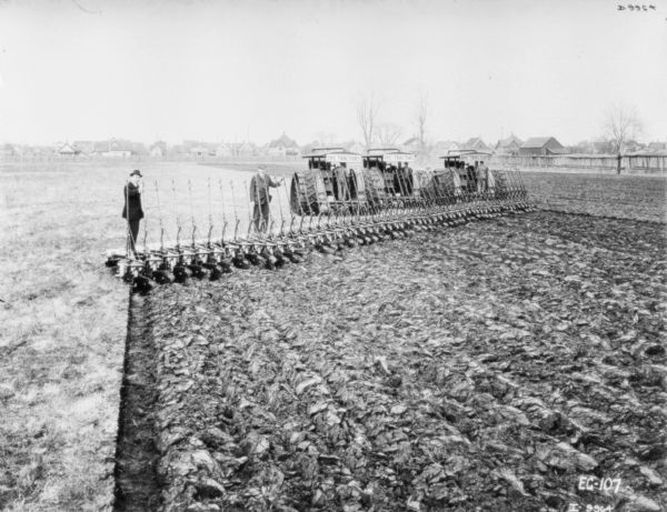 View of partially plowed field, with two men in suits standing posing on the left with 36 feet of plows, pulled by three tractors. The tractors each have a sign that reads: "IHC Tractors." The three men operating the tractors stand posing on the back of each tractor. In the background is a wood fence, and beyond are houses.