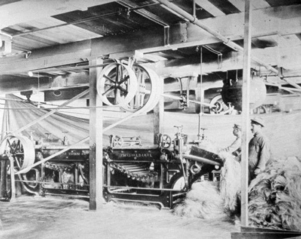 Spinning machine in factory. Belts attached to the ceiling power the machinery. Two men are standing on the right near bales of hemp. One of the men is feeding the hemp into the machine.