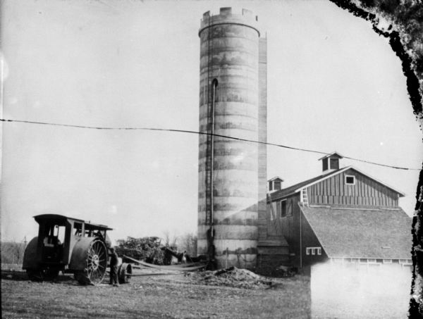 A man is standing near a tractor on the left, which is belt-driving an ensilage cutter near a silo. Farm buildings are on the right.