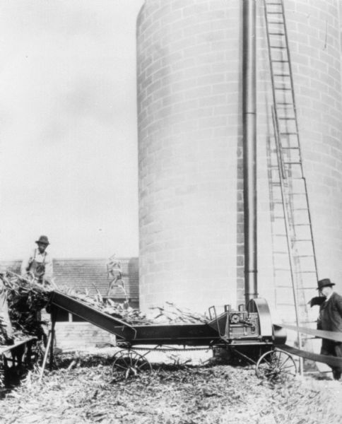 Two men are working with an ensilage cutter near a silo. The man on the left is feeding silage into the machinery. The man on the right is wearing a suit and hat, and is standing and watching near the ladder set up against the silo. The belts powering the ensilage cutter are on the far right. Farm buildings are in the background.