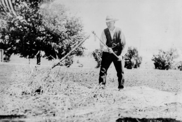 Man using a scythe in a field. In the background is a tree, and in the upper left corner is an American Flag.