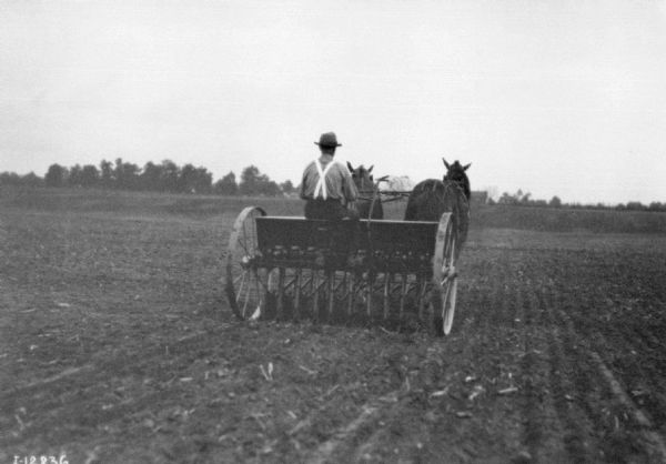 Rear view of a man using a horse-drawn seeder in a field. Trees and farm buildings are in the far background.
