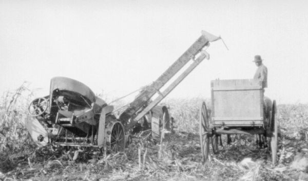 Rear view of tractor drawn corn picker. On the right is a man on a horse-drawn wagon.