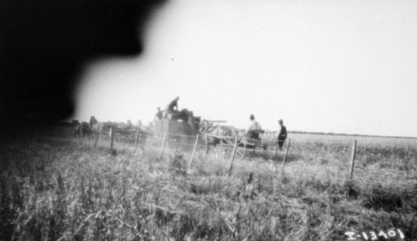 View across field and fence towards men using a (very early model) tractor to pull a binder. There are horses near the binder on the left, and a man is driving a horse-drawn buggy on the right.