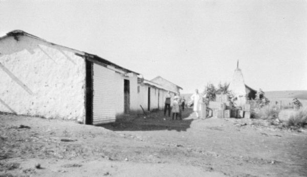 View across dirt yard towards a group of people posing outdoors near adobe houses which are in a row on the left.
