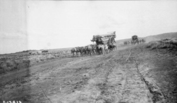 View across field towards a large team of horses pulling a tall wagon loaded with supplies on an unpaved road.