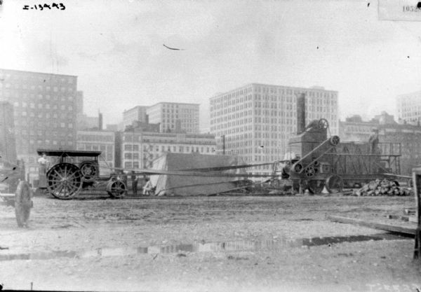 Tractor-powered rock crusher in what appears to be a factory yard. One man is standing on the tractor, and another man is standing on the rock crusher. Buildings are in the background.