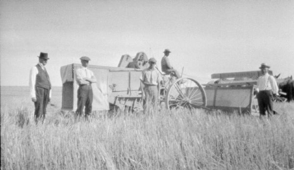 Four men are standing and posing in a field. Behind them a man is driving a horse-drawn Deering harvester thresher.