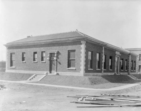 Exterior view of large, one-story brick building with tiled roof. Another building is in the background on the right. Lumber is piled in the yard in the right foreground.