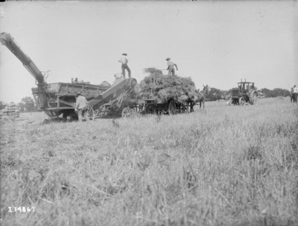 Men working with tractor powered thresher in a field. In the center a man stands on top of a horse-drawn wagon piled high with hay.