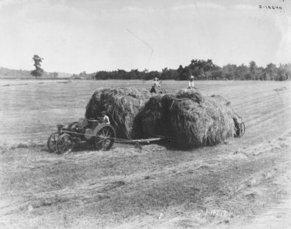 Elevated view of a tractor pulling two heavily loaded wagons in a field. Two men are standing on top of the wagons.