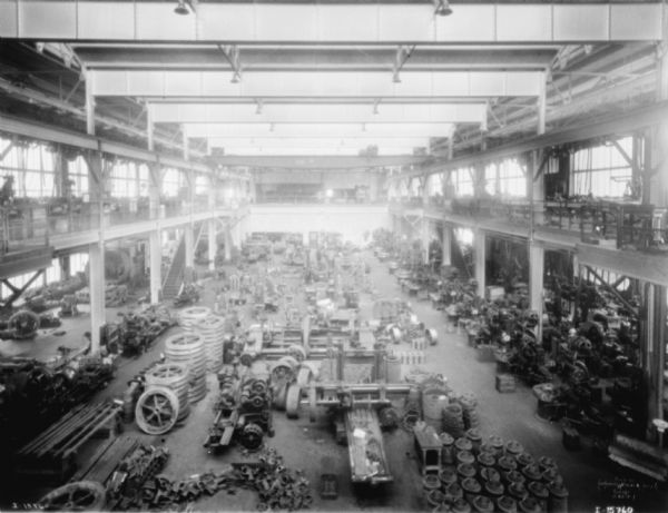 Elevated view of manufacturing area at McCormick Works. Parts are stacked on the ground floor. Upper level balconies are on the left and right. A crane is suspended on tracks high above the factory floor.