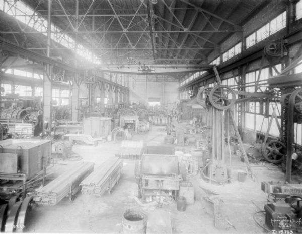 Elevated view of factory floor at McCormick Works. Parts and machinery are stacked and piled on the floor. A crane is suspended on tracks above the floor.
